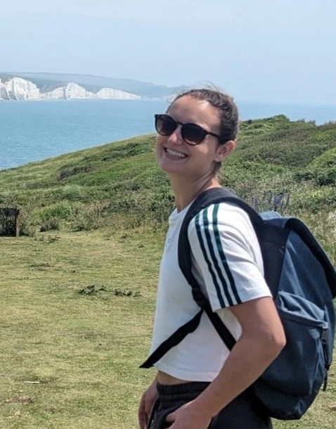 Jess McCormick, wearing a white shirt and sunglasses, smiling into the camera in front of a grassy landscape and ocean view