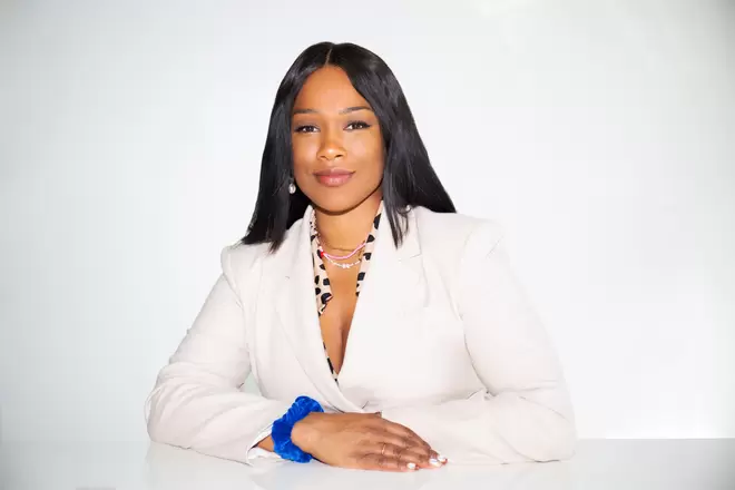 Sharmadean Reid MBE, founder of The Stack World, smiling at the camera, wearing a white suit and a blue scrunchie on her wrist.