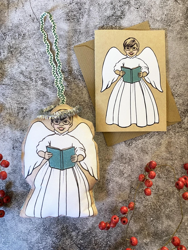 a hanging Christmas tree decoration of a boy angel carol singing next to a matching angel greeting card laid on a Kraft envelope