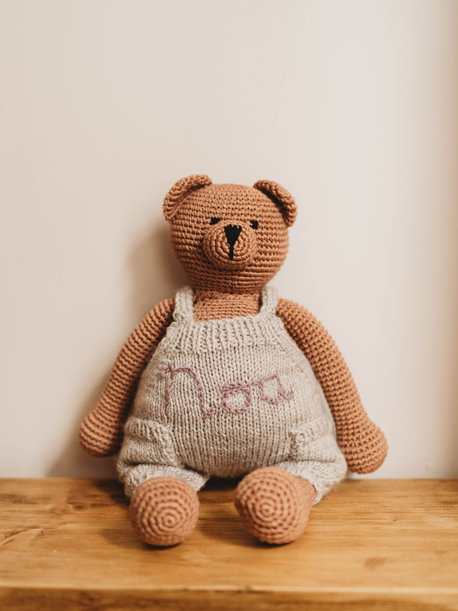 hand crochet brown teddy bear on wooden shelf  wearing beige hand knit personalised dungarees