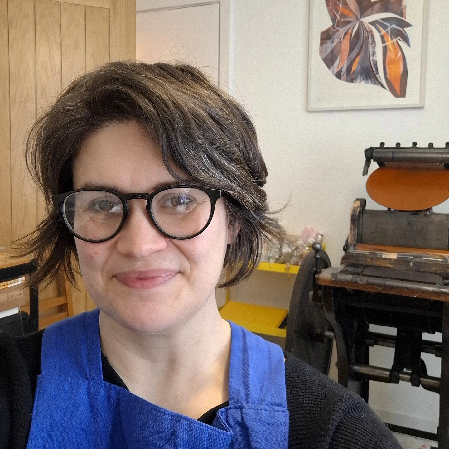 Anna Thundercliffe in her studio.  A woman smiles at the camera, there is a printing press and shelving behind her.
