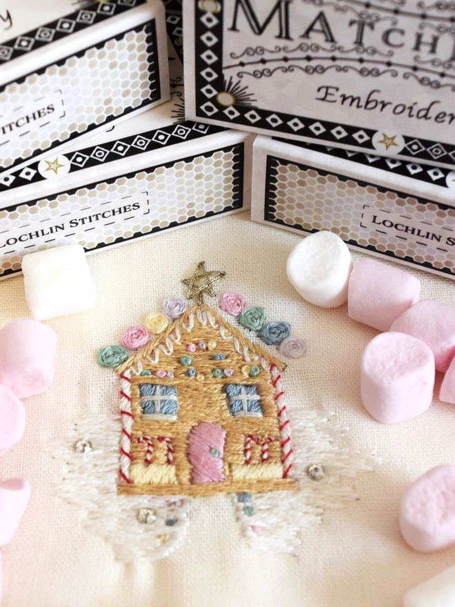 A delicious embroidered gingerbread house covered in sweets.  Displayed with mini marshmallows and matchbox packaging in the background.