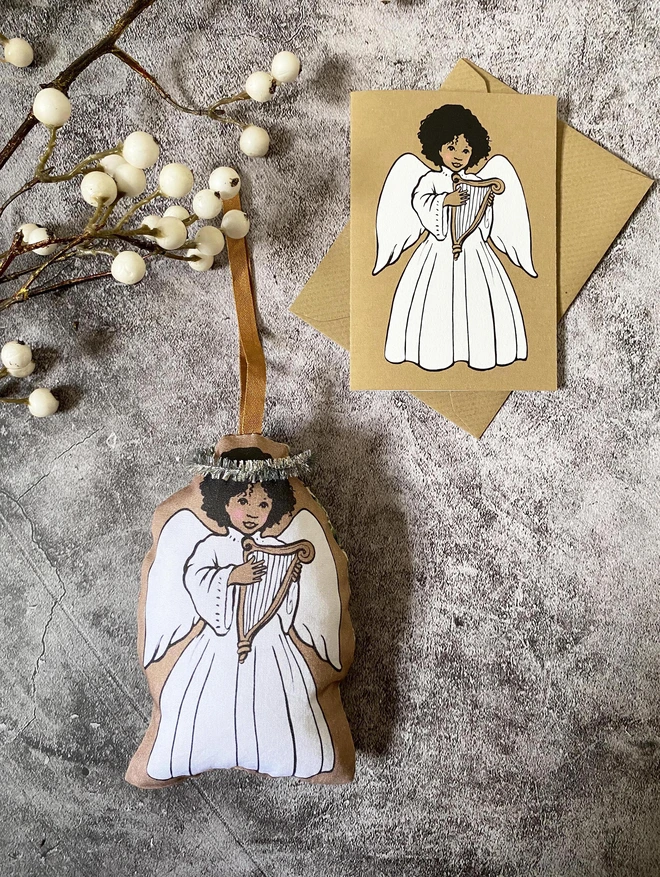 an angel Christmas tree hanging decoration made from fabric featuring an afro Caribbean girl holding a harp with a brown hanging ribbon laid next to a matching angel greeting card 