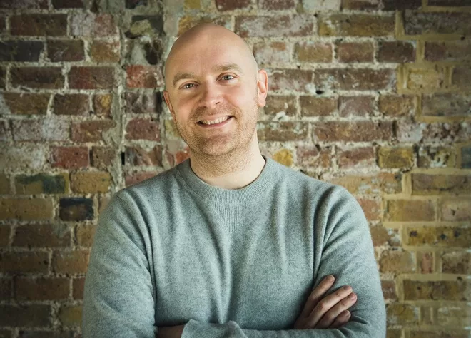 Will Beckett, co-founder of Hawksmoor, smiling at the camera, wearing a grey jumper, stood infront of a brick wall.