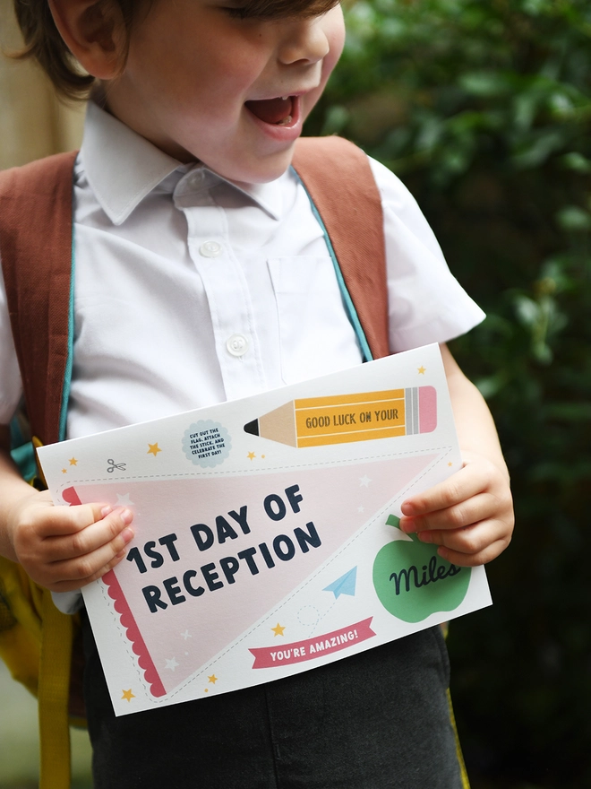 A young child, wearing a school uniform and backpack, holds a "1st Day of Reception" card with excitement. The card features encouraging messages and playful designs, perfect for celebrating a child's first day at school.