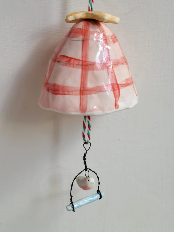 A hanging handmade bell in pink and red check and a tiny budgie figure hanging below