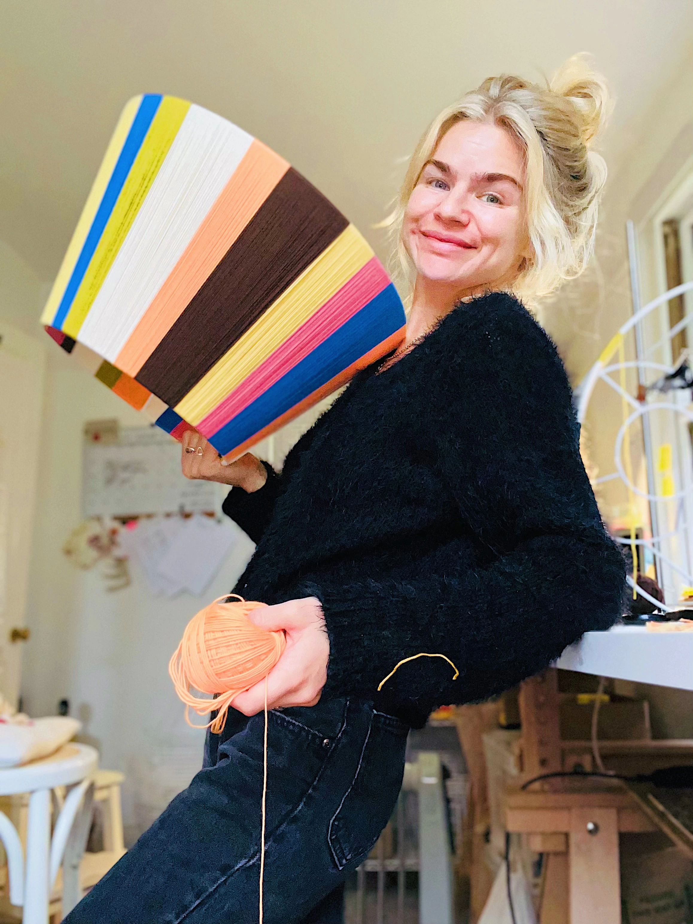 Sarah holding a lampshade in one hand and a ball of yarn in the other, leaning against desk. 