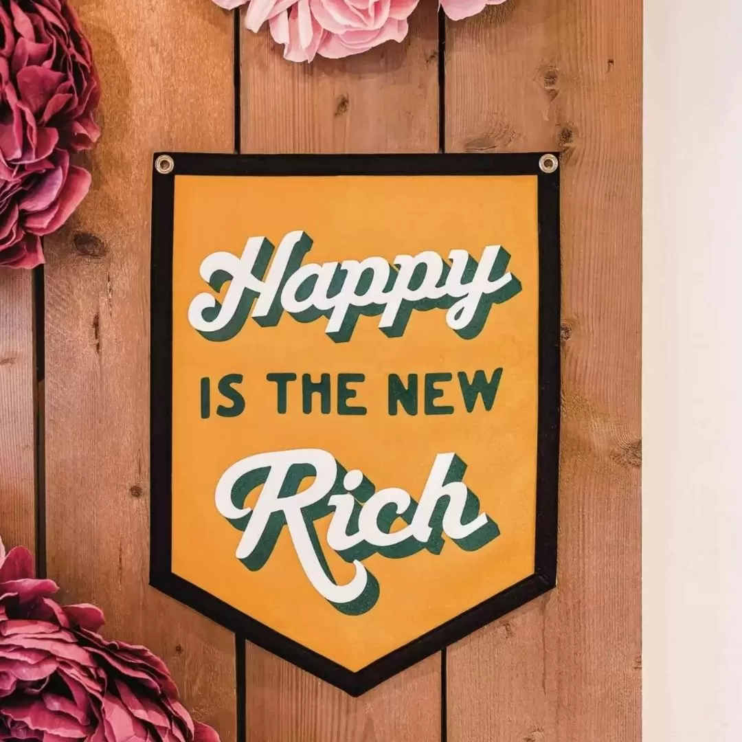 Happy is the new rich flag seen on a wooden wall with large flowers around it.