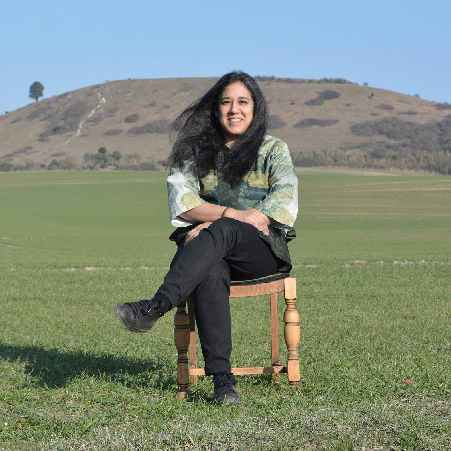 A profile picture of Natalie Thakur sitting on a chair in a green field with The Chiltern Hills in the background