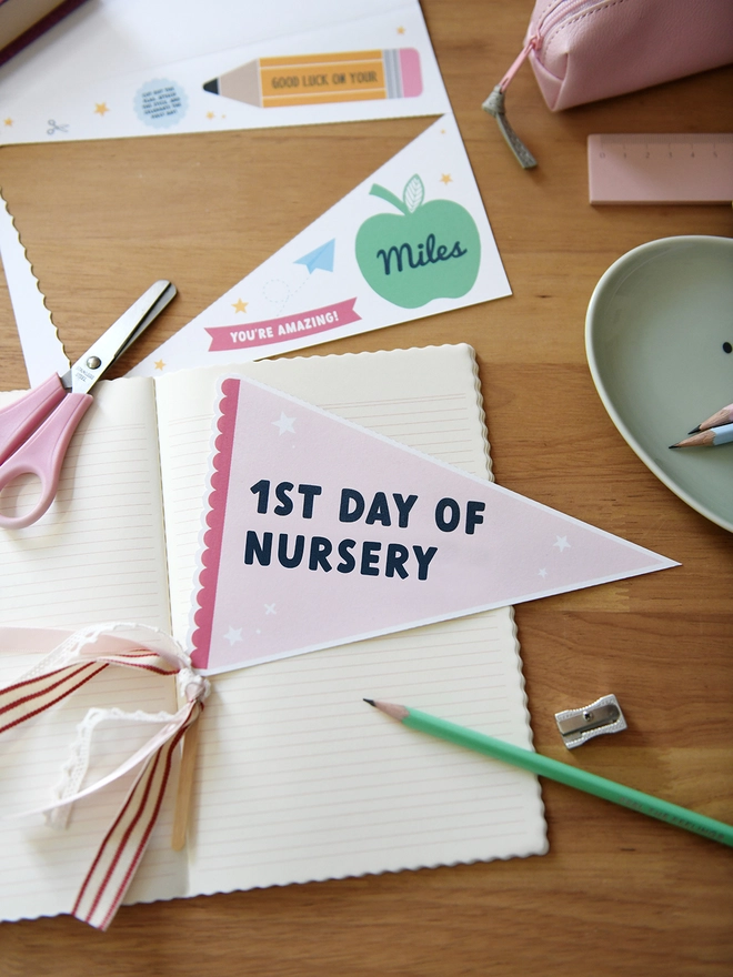 A pink pennant flag with the words "1st Day Of Nursery" lays on an open notebook on a wooden desk. Various stationery items surround it.