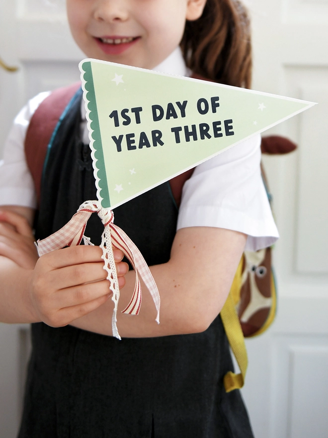 A child in a school uniform holds a green pennant flag that reads "1st Day of Year Three," adorned with ribbons.
