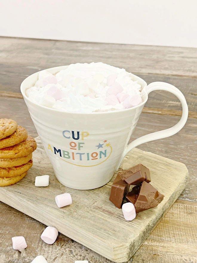 a porcelain large fluted mug with 'Cup of Ambition' printed on the outside in tones of blue, red and greens. sits on a wooden board with a pile of chocolate chunks and a stack of cookies. The mug is filled with a decadent hot chocolate complete with whipped cream and marshmallows