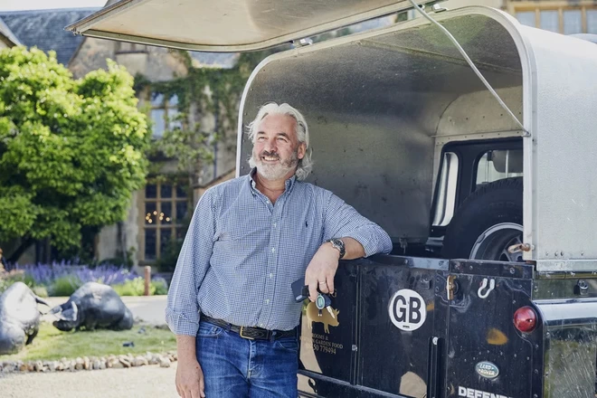 Robin Hutson, founder of The Pig Hotels, smiling into the distance, stood next to his landrover in a blue shirt.