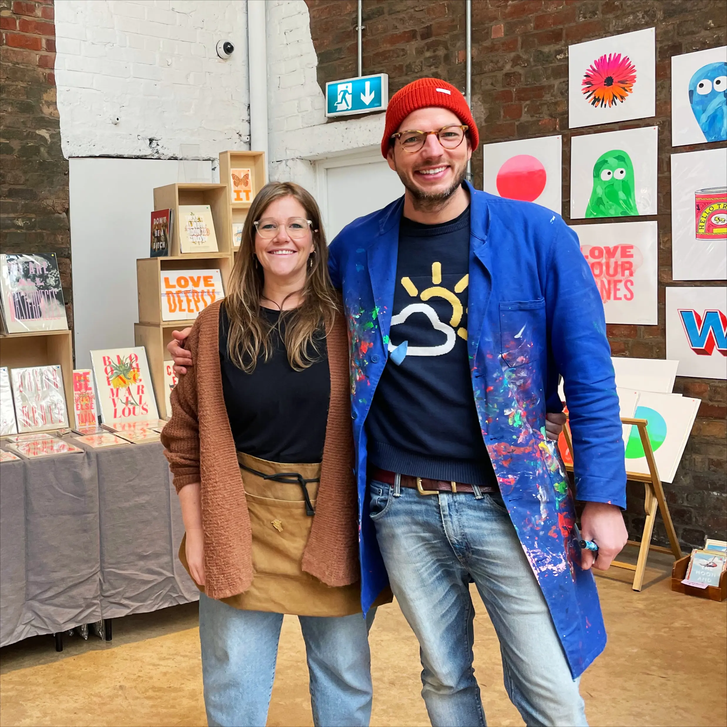 A photograph of Matt and Lucy, the founders of Basil & Ford. They are standing infront of a market stall featuring all their bright flouro screenprints. 