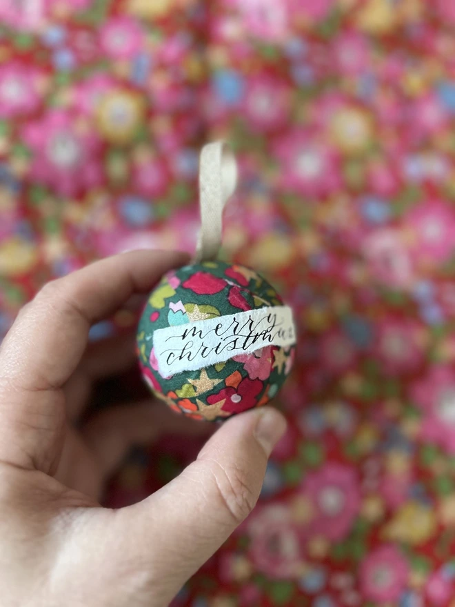 Mini Liberty fabric Christmas bauble with Merry Christmas label held in front of a Liberty fabric backdrop 