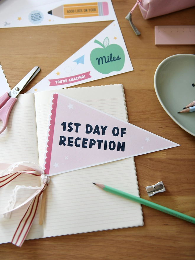 A pink pennant flag with the words "1st Day Of Reception" lays on an open notebook on a wooden desk. Various stationery items surround it.