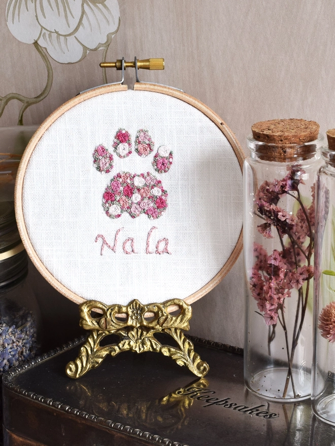 An embroidered Pink Roses Cats Paw, of woven wheel roses in 5 shades of pink with French Knot green grass.  Displayed in a hoop frame on a gold metal stand on top of a silver keepsake box.