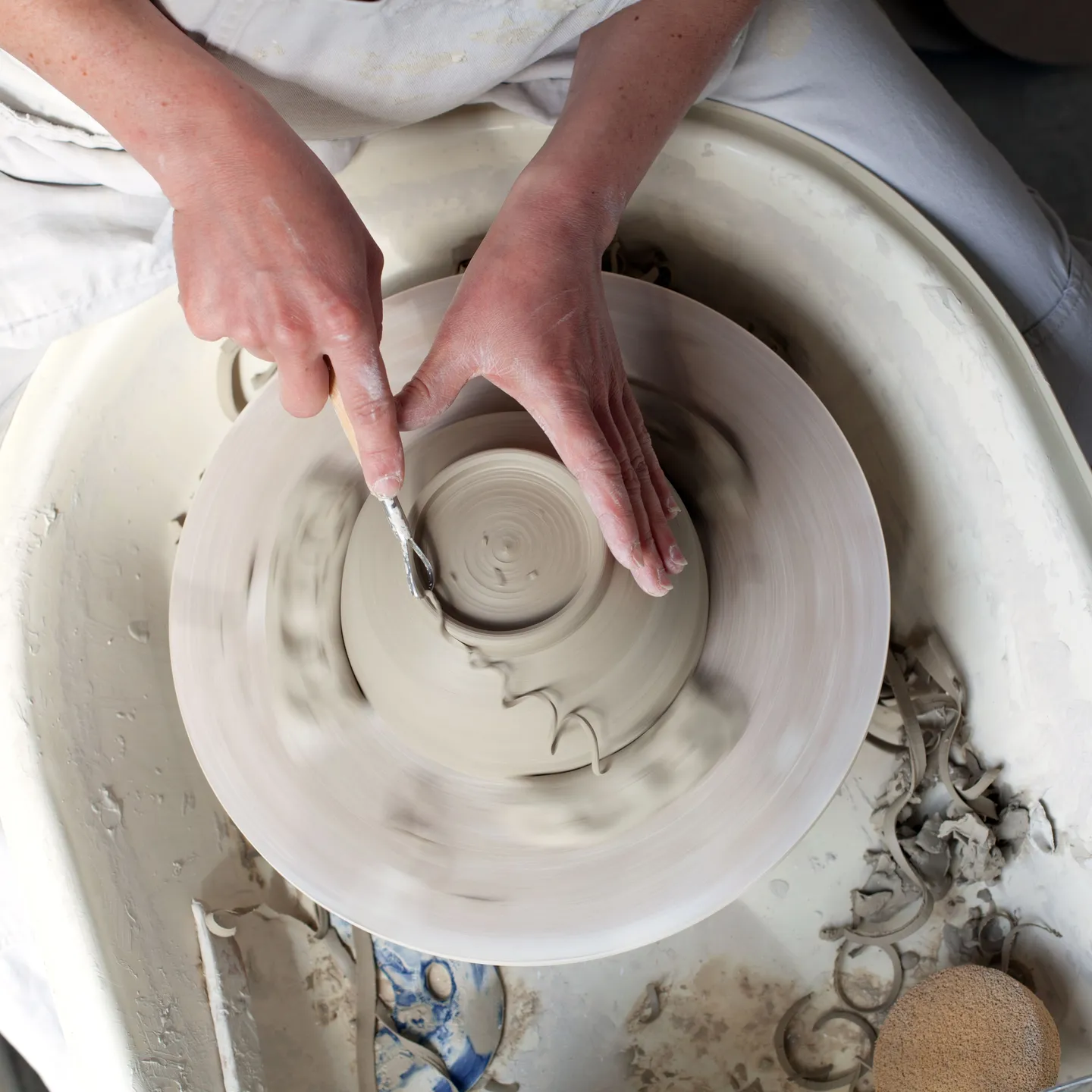 Looking down on a female hand wrapped around an upside down bowl on a pottery wheel. The other hand holding a tool with a curl of clay coming from it and from the footring of the bowl. 
