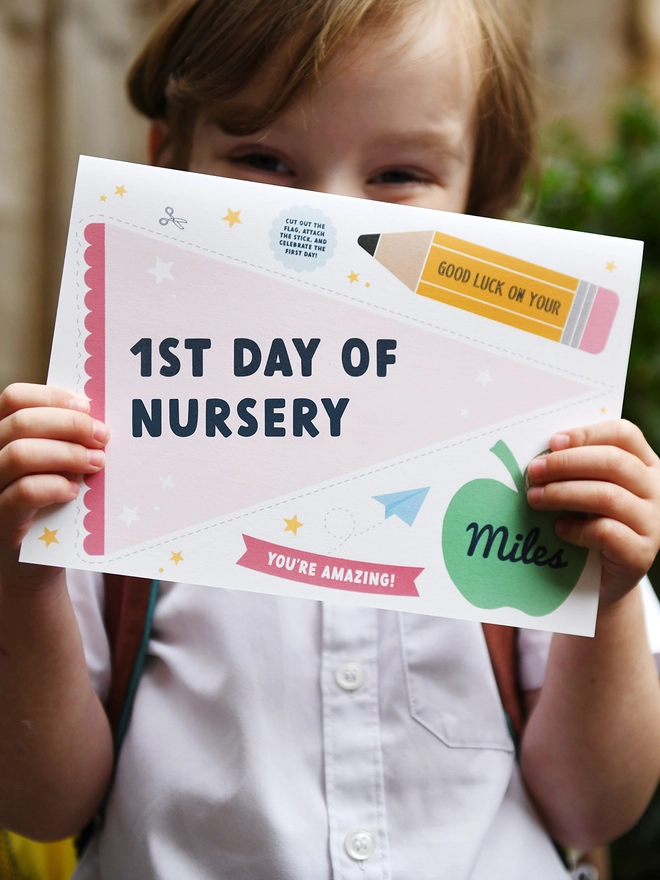 A young child, wearing a school uniform and backpack, holds a "1st Day of Nursery" card with excitement. The card features encouraging messages and playful designs, perfect for celebrating a child's first day at school.