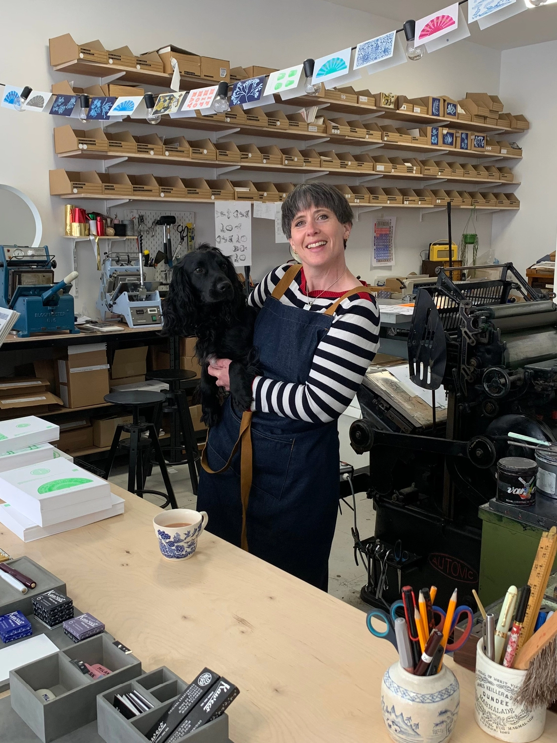 A smiling women stands in a colourful shop front holding a small black dog.