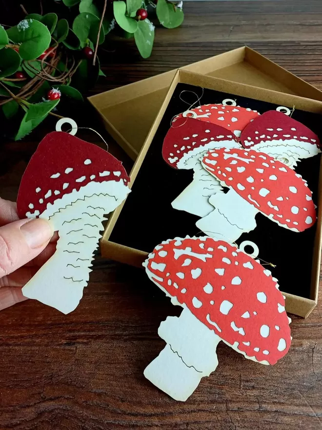 A mix of 6 red and white paper mushroom hanging decorations are displayed in a kraft brown gift box with black interior.  One of the decorations is being lifted out of the box and held in a woman's hand.