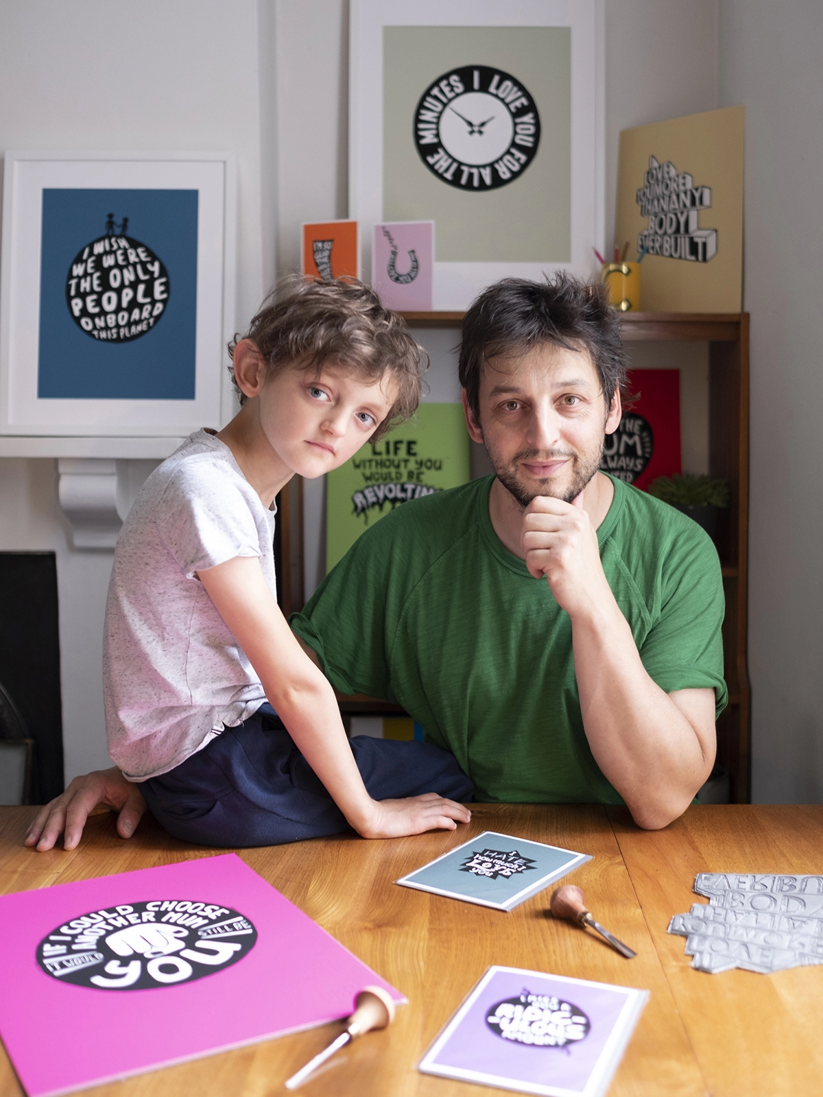 father and autistic son sitting at a table surrounded by linocut art prints of phrases said by the son and illustrated by his Dad