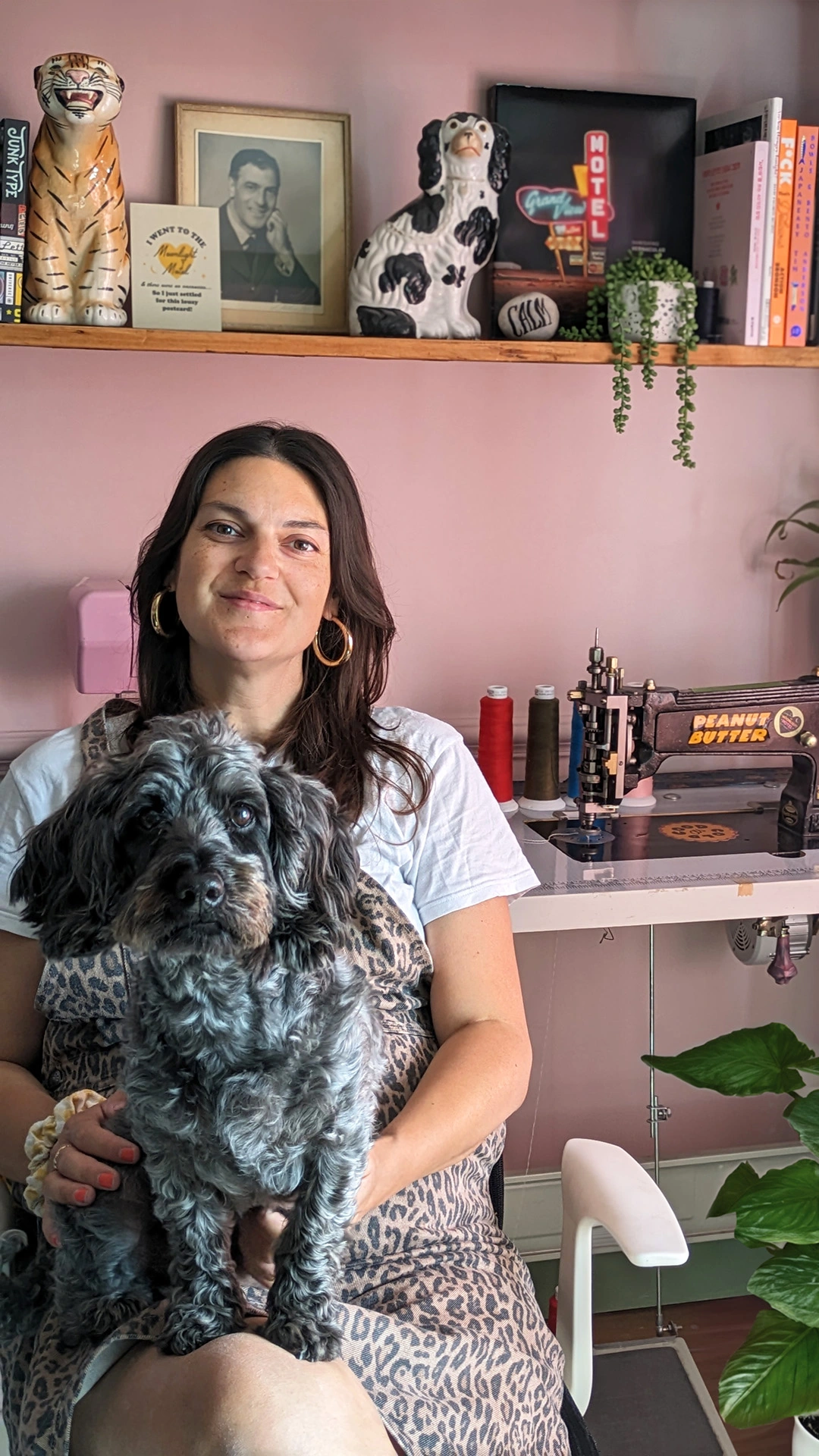 Lana & her dog Sumo sitting in front of her Chainstitch Machine in her workspace, surrounded by books, plants, pictures & dog sculptures