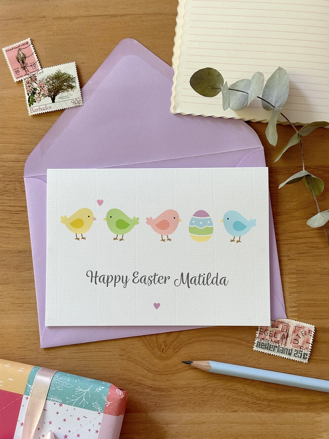 A personalised Easter card with pastel illustrations of spring chicks lays on a purple envelope, on a wooden desk.