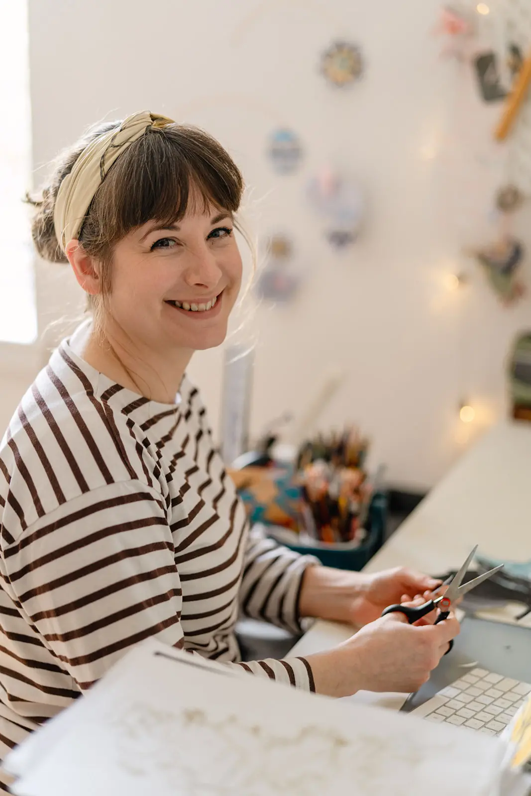 Snmiling woman in striped top and headband sits at desk, holding scissors