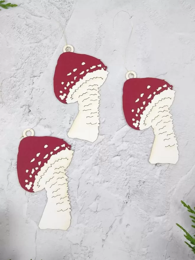 Three dark red and white paper mushroom decorations lay flat on a pale grey surface. The mushrooms have dark red  pointed caps with white spots and have thin gold thread hanging loops.