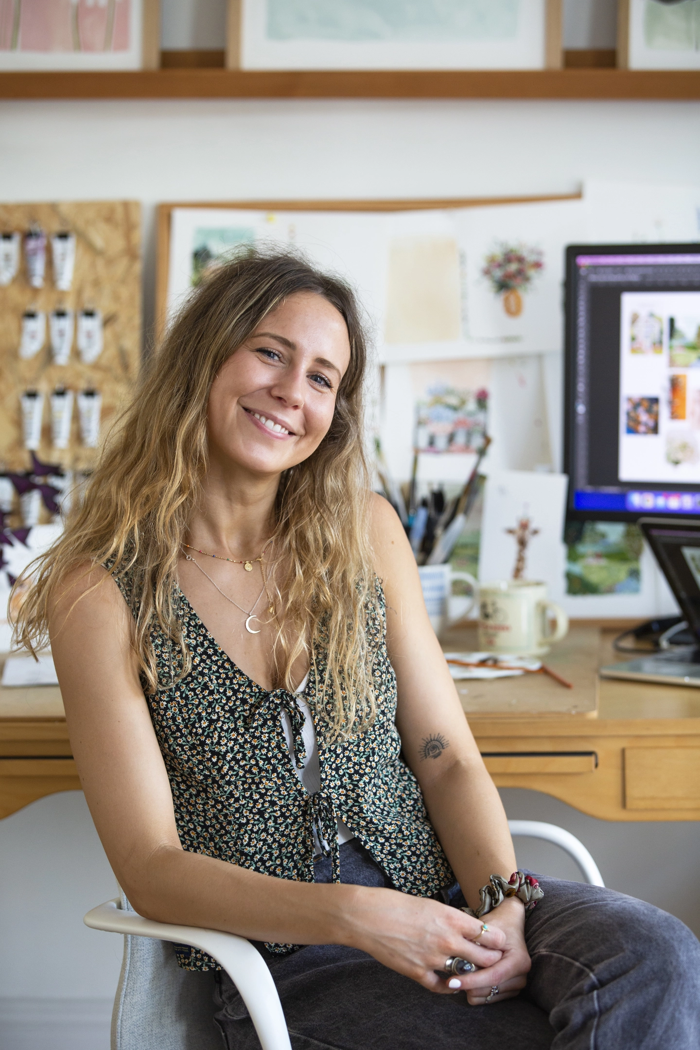 A photo of designer Louise Mulgrew sat at her desk in her art studio smiling facing the camera. Behind her is a noticeboard of her paintings and illustrations and her desk full of gouache paints and art materials.