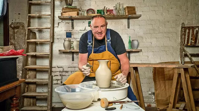 Keith Brymer Jones, British Designer, Master Potter & Author, smiling at the camera, wearing a mustard apron while building a clay pot.