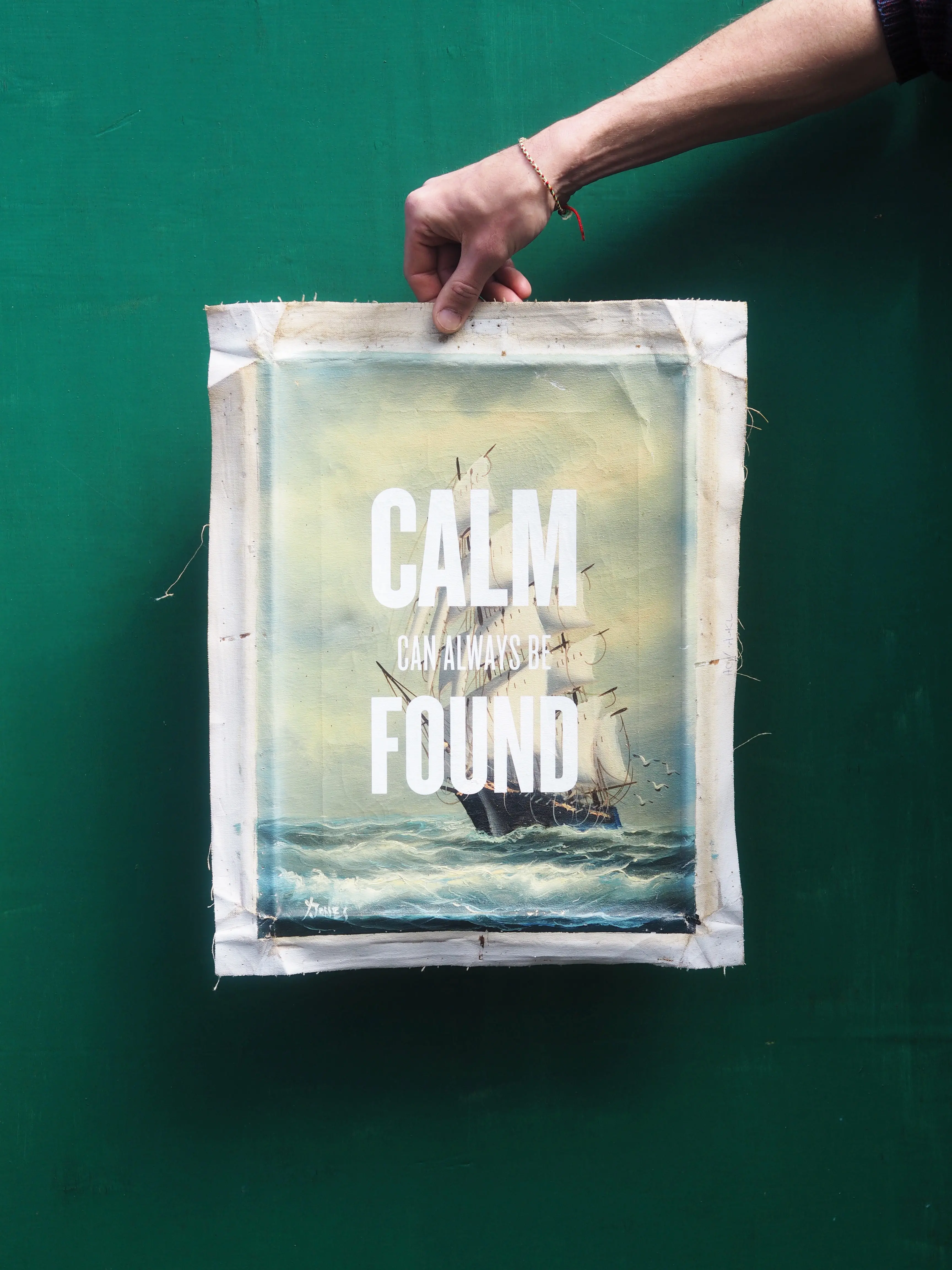 An old oil canvas is being held up by one hand infront of a dark green background. The oil painting features an image of a large sail ship sailing on the sea. Over the top in white typography are the words 'calm can always be found'