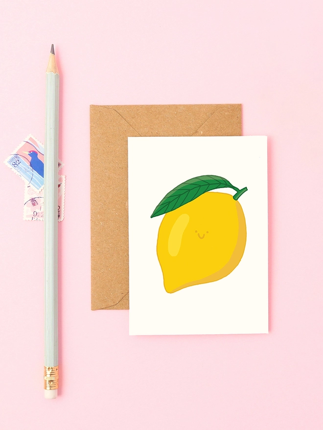 Mini lemon greeting card styled with kraft envelope, mint pencil and pink background, featuring a smiling yellow lemon with a green leaf illustration
