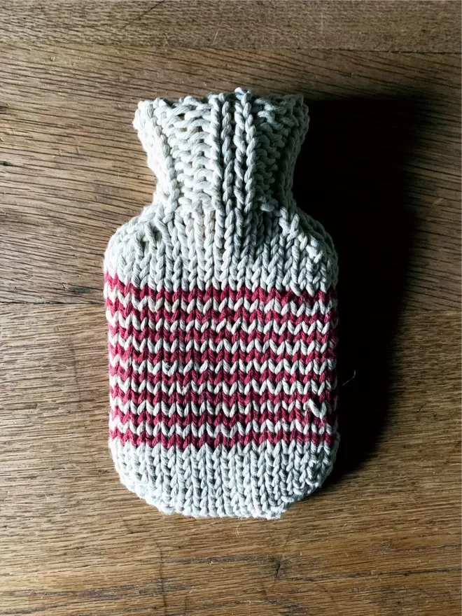 Mini Hot Water Bottle in hand knit string cover with red stripe on an oak table