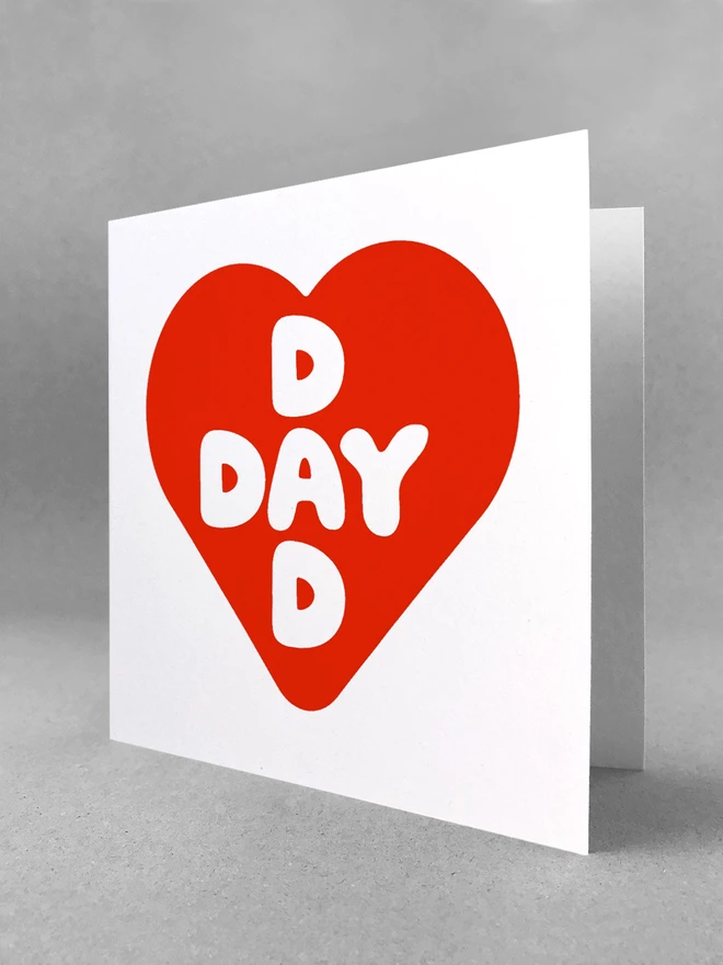 Red screenprinted heart contains intersecting words Dad and Day, a square card stood in a white studio backdrop slightly open angle.