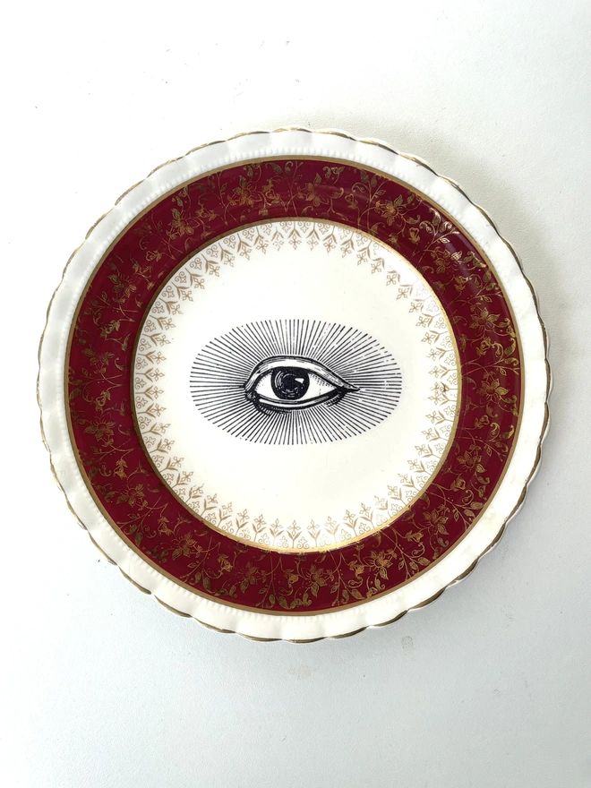 a really ornate red, gold and white china vintage plate has the vintage black and white image of an eye in the centre