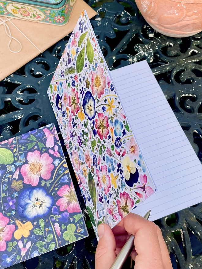 Hand Opening Floral Notebook, Revealing Lined Pages, Smaller Notebook from Same Range Nearby, on Garden Table
