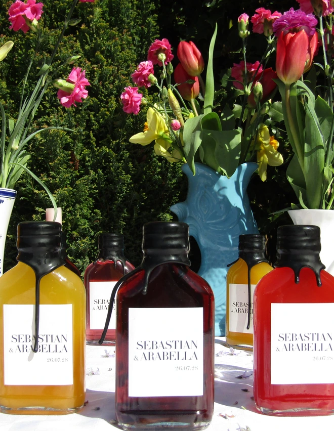 multiple glass bottles of various coloured cocktails with white label with names of wedding couple, against floral backdrop