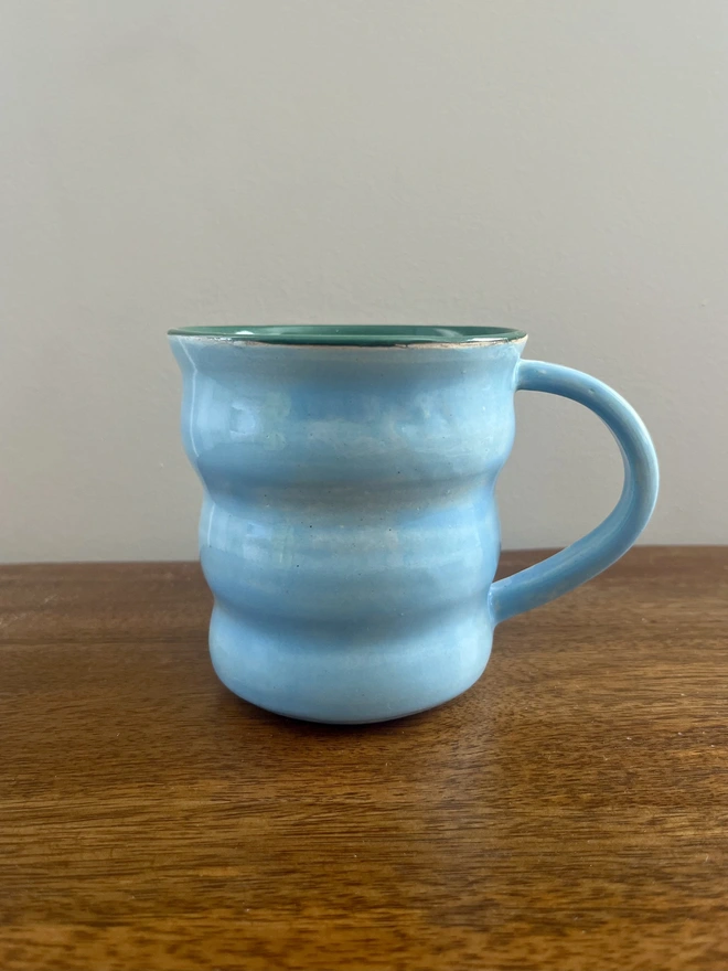 Hand-thrown 400 ml ceramic coffee mug with a wavy shape, glossy sky blue glaze, forest green interior and rounded handle, handmade by Hannah Inskip Pottery. Mug is sitting on a wooden table.