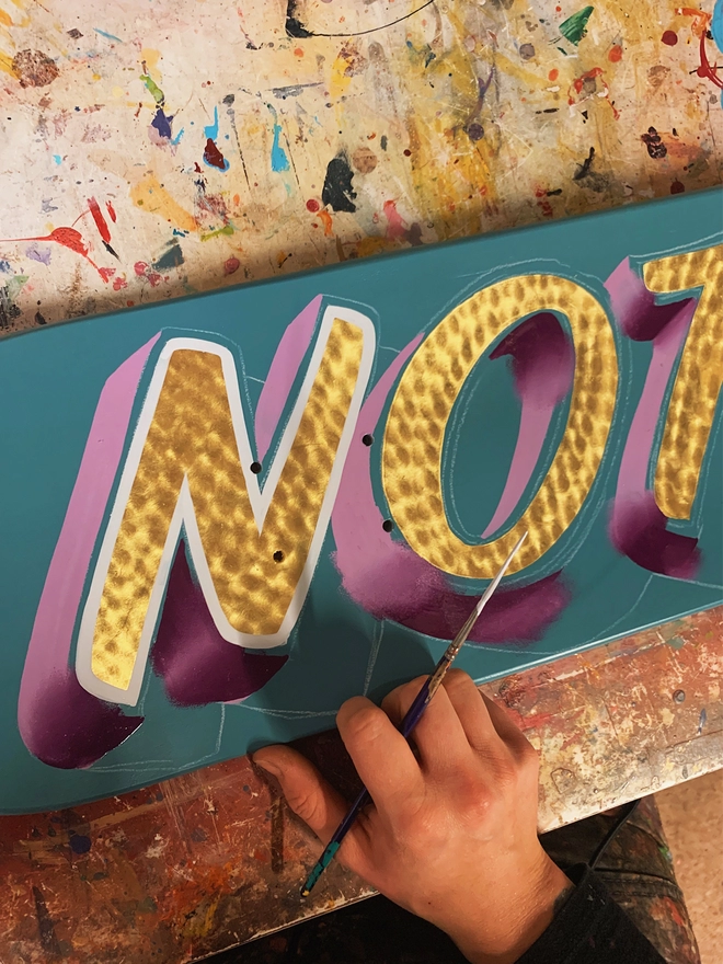 Work in progress shot a spun gold lettering spelling 'NOTTS' on a turquise deck with purple and white shade and outline.