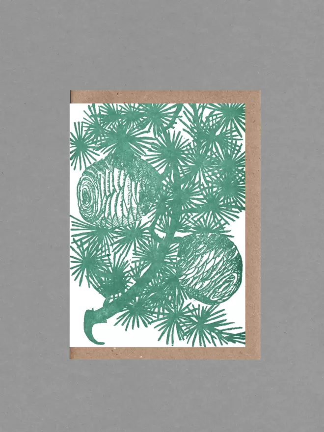 Greetings card with an illustration of a cedar branch in green. Behind the card is a brown envelope on a grey backround.