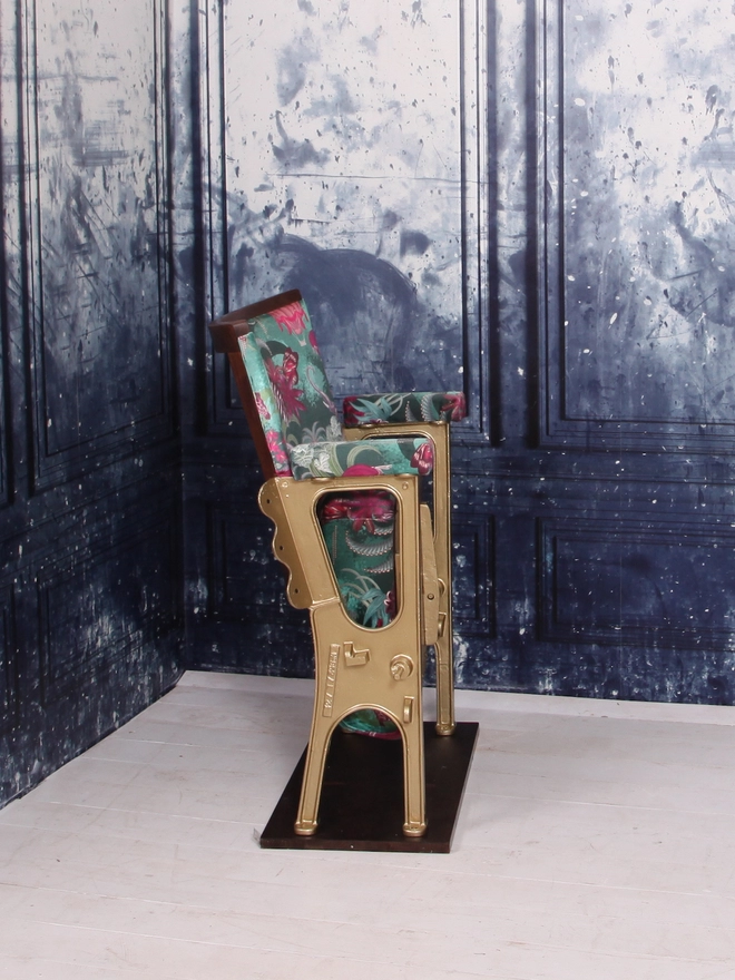 Side view of a single vintage cinema seat upholstered in a green and pink safari velvet, against a blue marbled wall.  The seat of the cinema seat is up