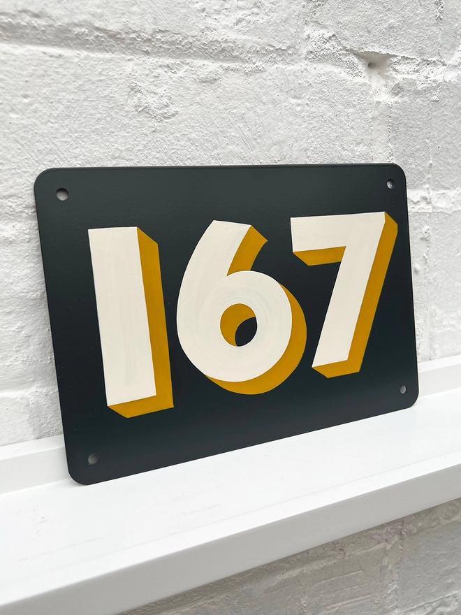 Hand painted off white and mustard house number 167 on an anthracite grey metal plaque against a white brick wall. 
