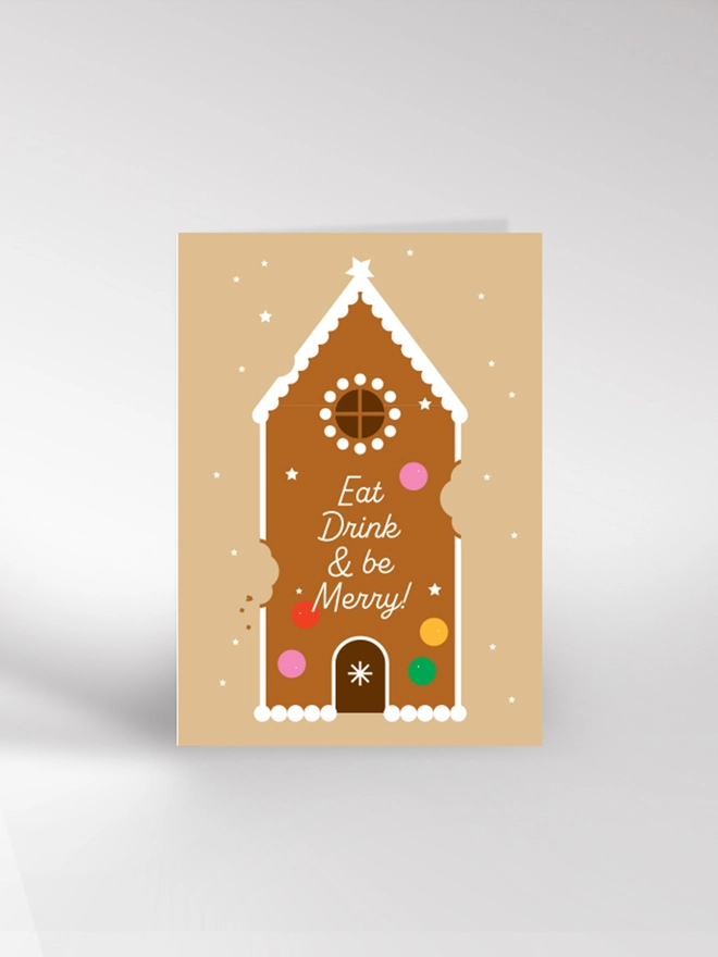 Contemporary illustrated Christmas card showing a gingerbread house with the words 'Eat, Drink & Be Merry' piped in white icing 