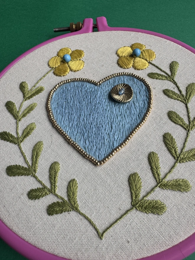 A close up of an embroidery hoop containing a hand stitched blue heart edged in gold beads with yellow flowers on long green stalks either side