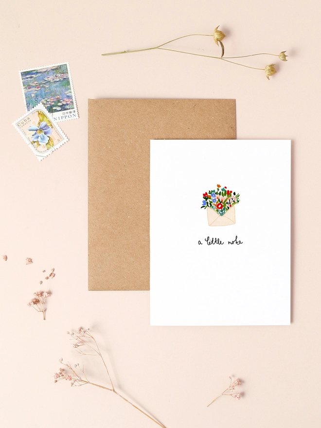 An image of a mini, rectangular white greetings card with an illustration of a small envelope bursting open that's been filled with colourful flowers. A hand written 'a little note' message is seen under the central illustration.