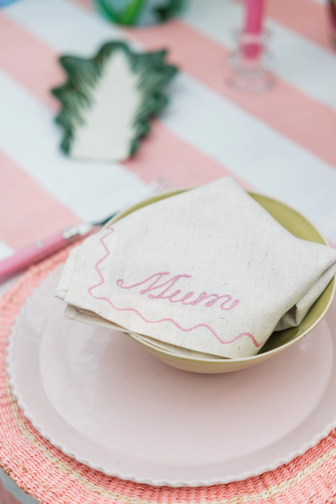 Personalised linen napkins with pink, 'Mum' folded into a tablescape with pink and peach placemates and plates on a pink and white striped tablecloth