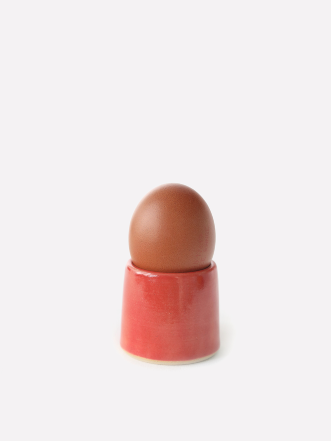 A handmade ceramic egg cup in cherry red glaze holding a brown egg, photographed against a plain white background