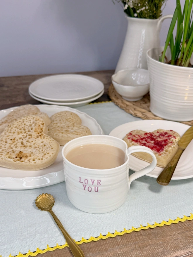 hand thrown porcelain cream mug with pink lustre text on the side that says LOVE YOU. The mug is sat upon a sage table cloth amongst a tea time scene of heart shaped crumpets